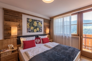 A bedroom with a wooden headboard, a large bed with red and white bedding, a nightstand with a lamp, and a large window with a view of a forested area.