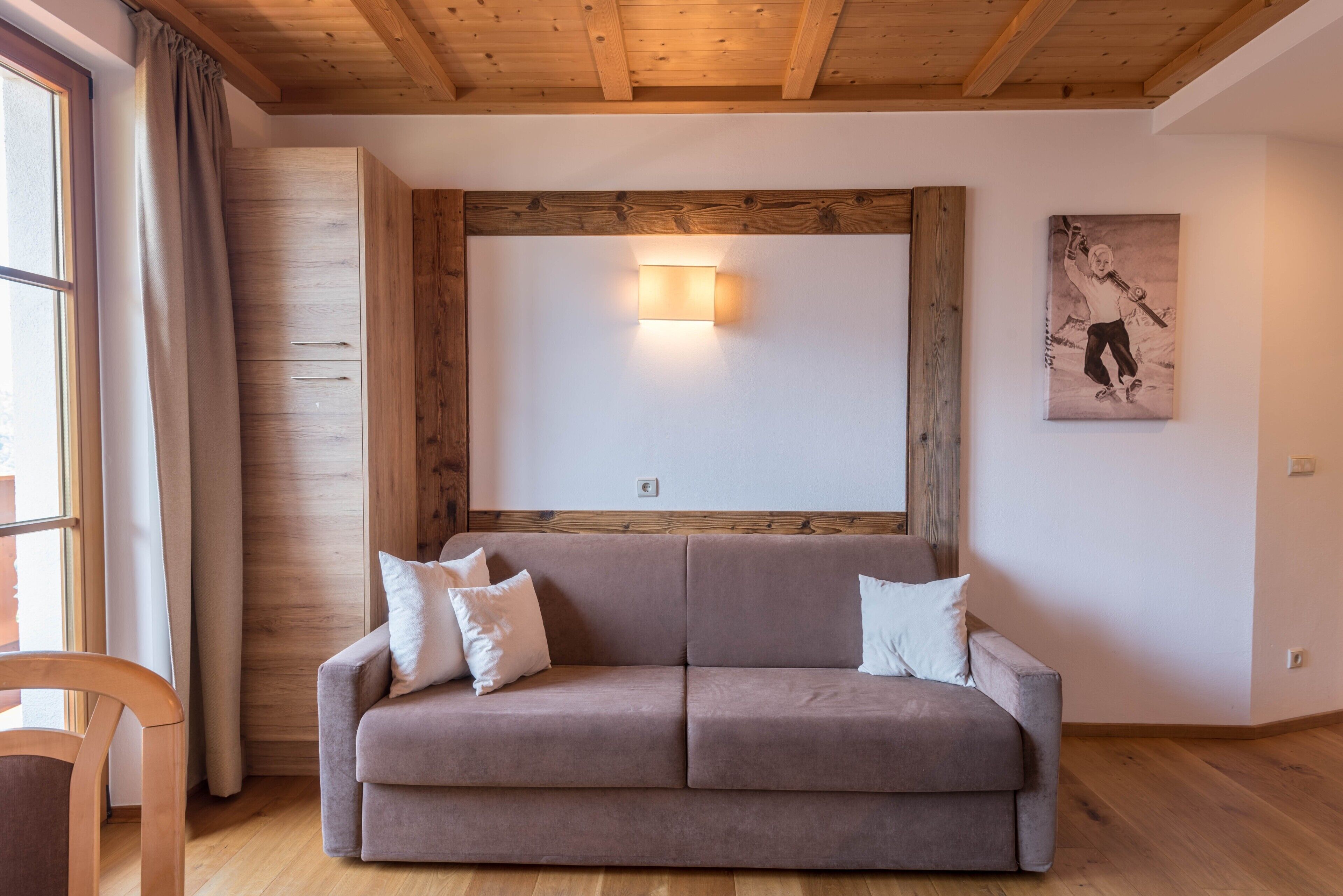 Ein Zimmer mit Holzboden, einem beigefarbenen Sofa mit Kissen, einem Holzschrank, einer Wandleuchte und einem gerahmten Bild.