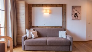 A room with a wooden ceiling, a beige sofa with pillows, a wooden cabinet, a wall-mounted light, and a framed picture.