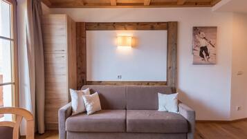 Ein Zimmer mit Holzboden, einem beigefarbenen Sofa mit Kissen, einem Holzschrank, einer Wandleuchte und einem gerahmten Bild.