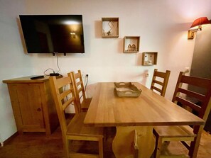Un comedor con una mesa de madera, sillas, un televisor montado en la pared y un pequeño estante con adornos.
