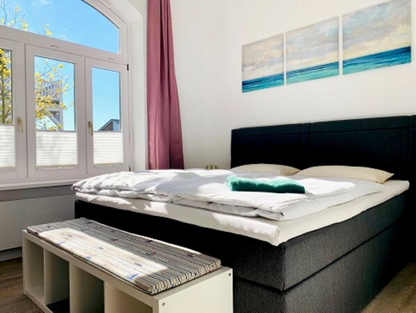 A bedroom with a bed, a window with blinds, a bedside table, and a wall with three paintings.