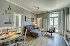 Una sala de estar moderna con un sofá gris, una mesa de comedor preparada para dos personas y un ventanal con vistas a edificios.