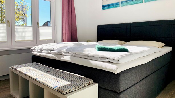 Un dormitorio con cama, una banca, ventana con persianas, cortina y un muro con pinturas.