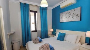 A hotel room with a bed, two bedside tables, a lamp, a trash bin, a window with curtains, and a wall-mounted air conditioner. - Casa Carra - Island Escape by Villas Now (Playa Blanca)
