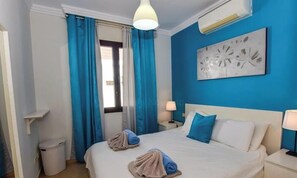 A hotel room with a bed, two pillows, a nightstand, a lamp, a trash bin, and a window with curtains.