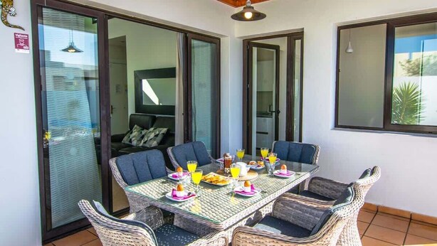 A dining area with a glass table, wicker chairs, and a view through the glass doors. - Casa Carra - Island Escape by Villas Now (Playa Blanca)