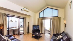 A spacious living room with a large TV, a sofa, a coffee table, and a balcony with sliding glass doors. - Casa Carra - Island Escape by Villas Now (Playa Blanca)