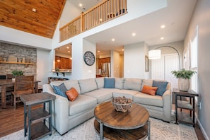 A spacious living room with a wooden ceiling, a stone wall, a dining area, and a kitchen in the background.