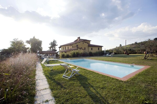A large house with a swimming pool, surrounded by a well-maintained lawn and a few lounge chairs.