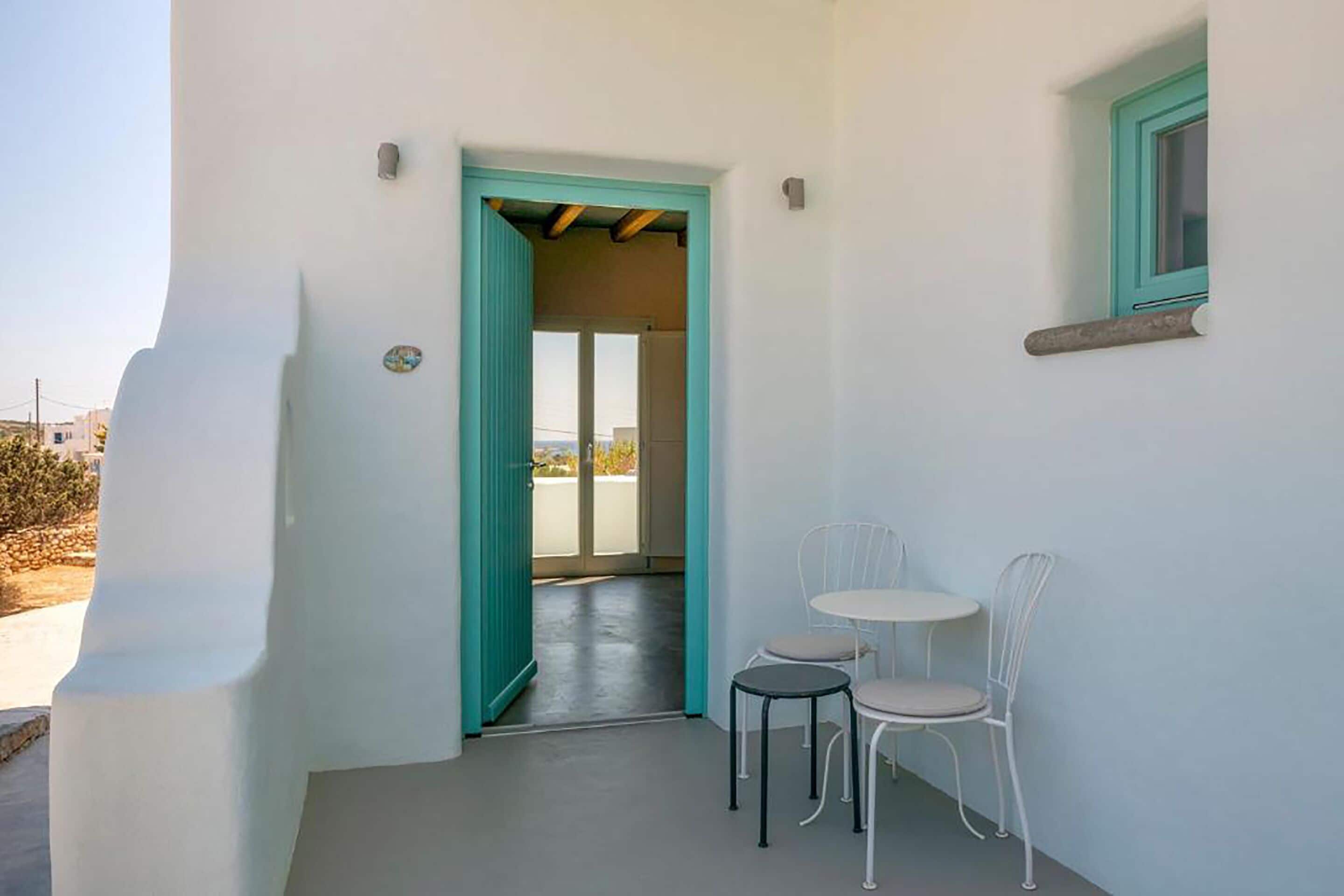 A narrow hallway with a teal door leading to a balcony. There are two white metal chairs and a small round table on the balcony.