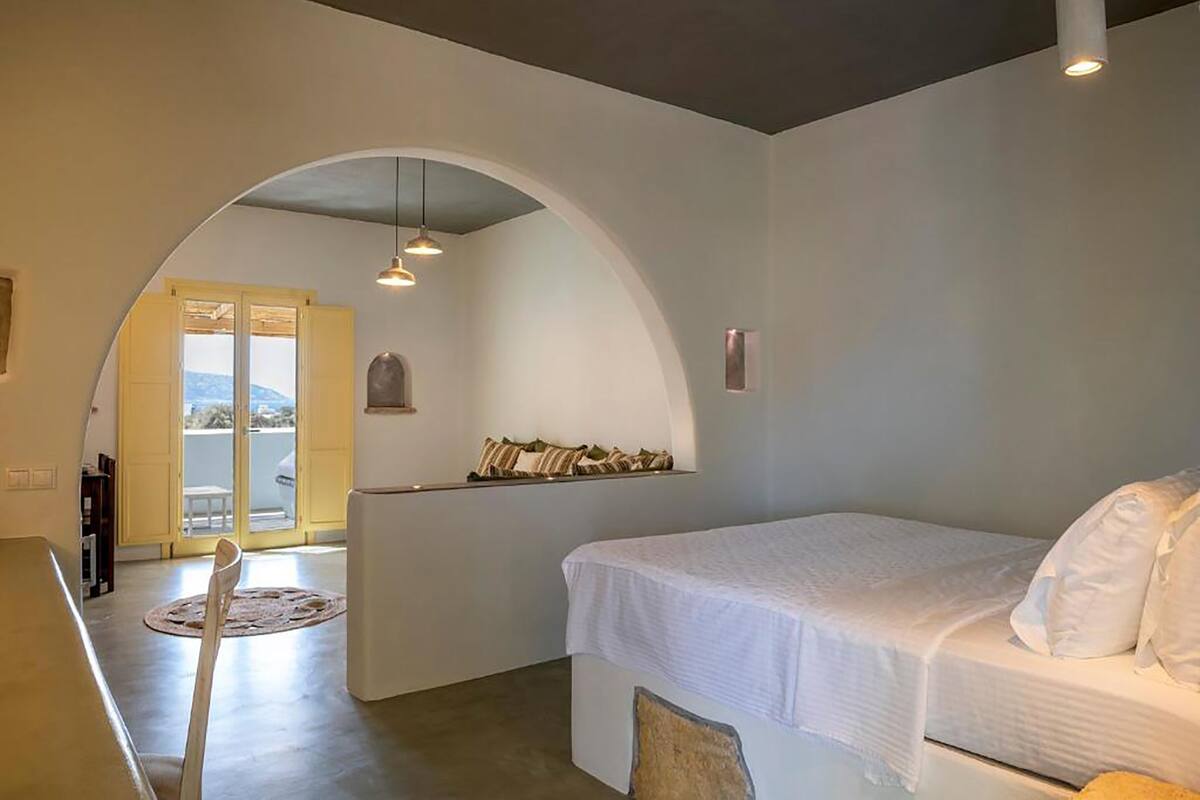 A bedroom with a bed, a small seating area, a window, and a door leading to an outdoor balcony.
