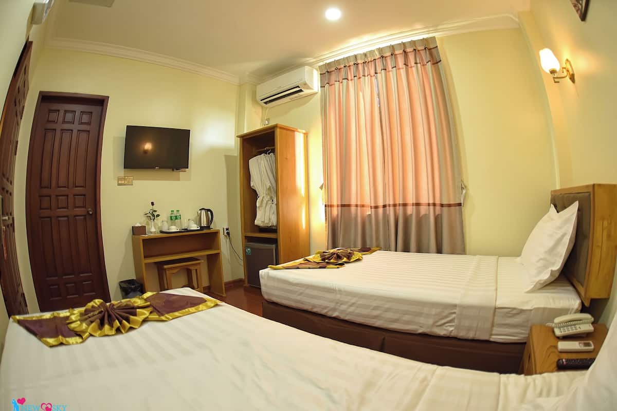 A hotel room with two beds, a TV, an air conditioner, a wardrobe, and a desk with a kettle and a rose.