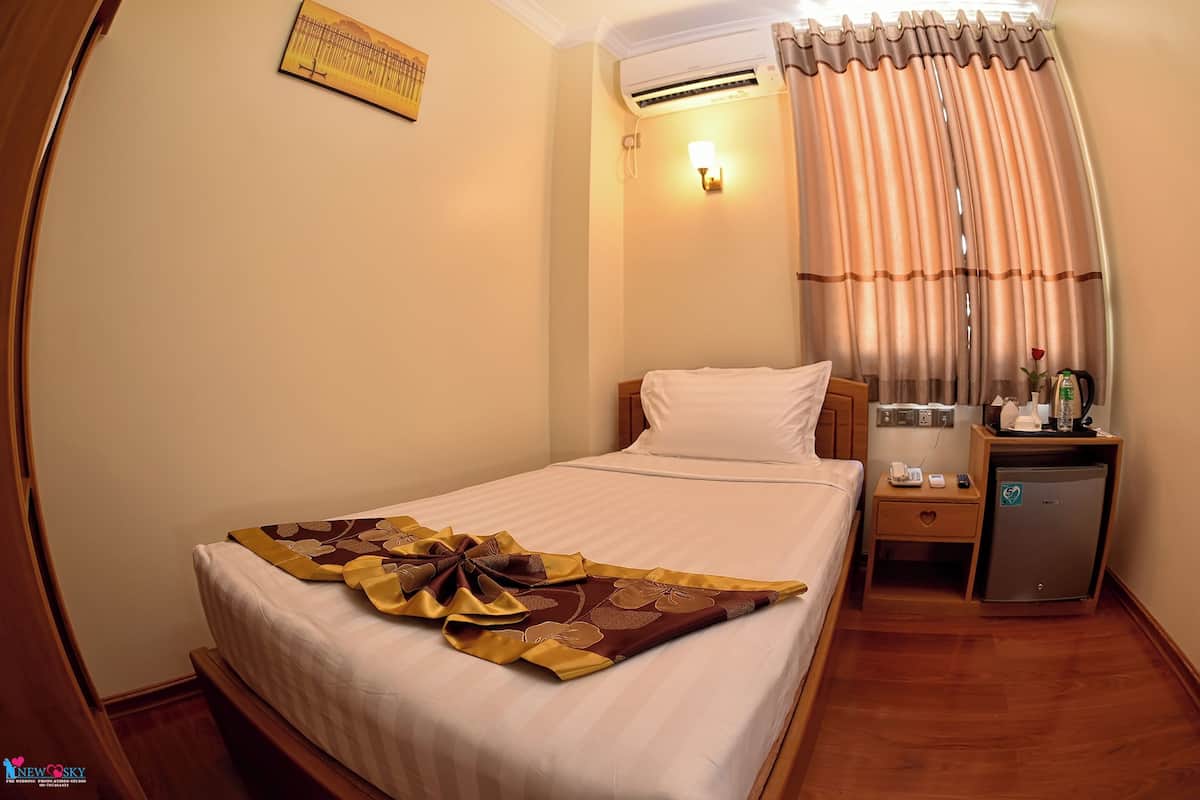 A hotel room with a neatly made bed, a bedside table with a lamp, a small refrigerator, and a window with curtains.
