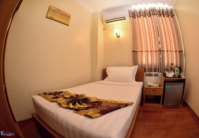 Habitación de hotel con una cama bien hecha, una mesita de noche con lámpara, un refrigerador pequeño y una ventana con cortinas.