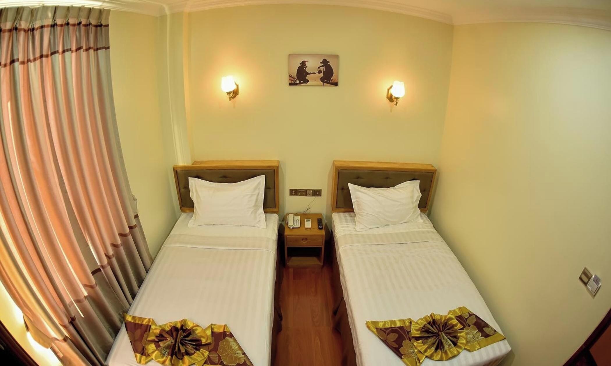 Habitación de hotel con dos camas individuales, una mesita de noche con teléfono y control remoto, y una lámpara de pared.