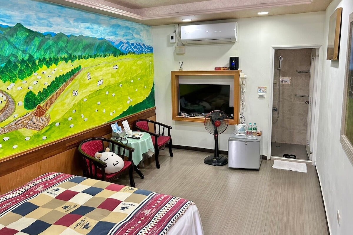 Habitación de hotel con un mural de un paisaje verde, una cama, un escritorio con una silla, un televisor, aire acondicionado y ducha.