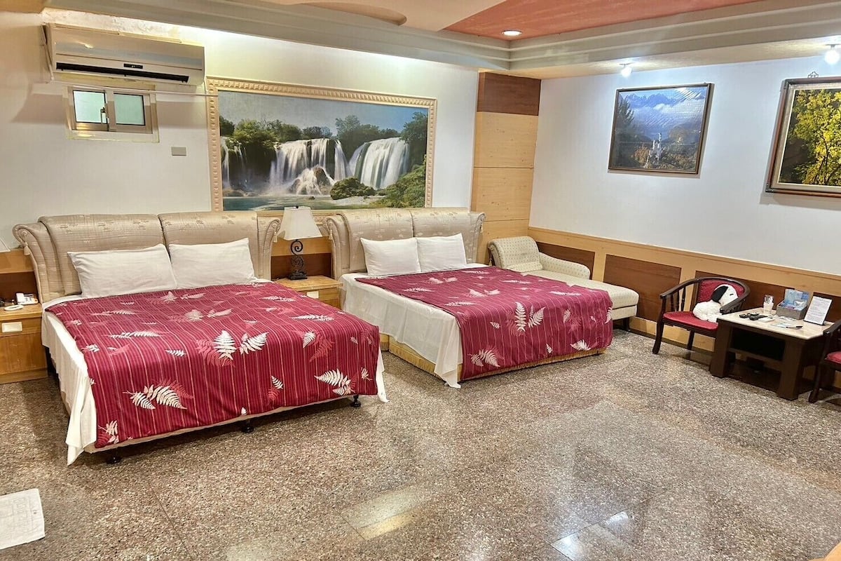 Habitación de hotel con dos camas, un cuadro de una cascada, una mesita con una silla y una ventana con vista a árboles.