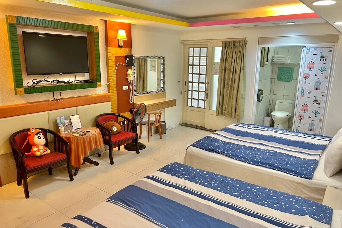 Habitación de hotel con dos camas, televisión, escritorio con silla, un oso de peluche y baño visible a través de la puerta.