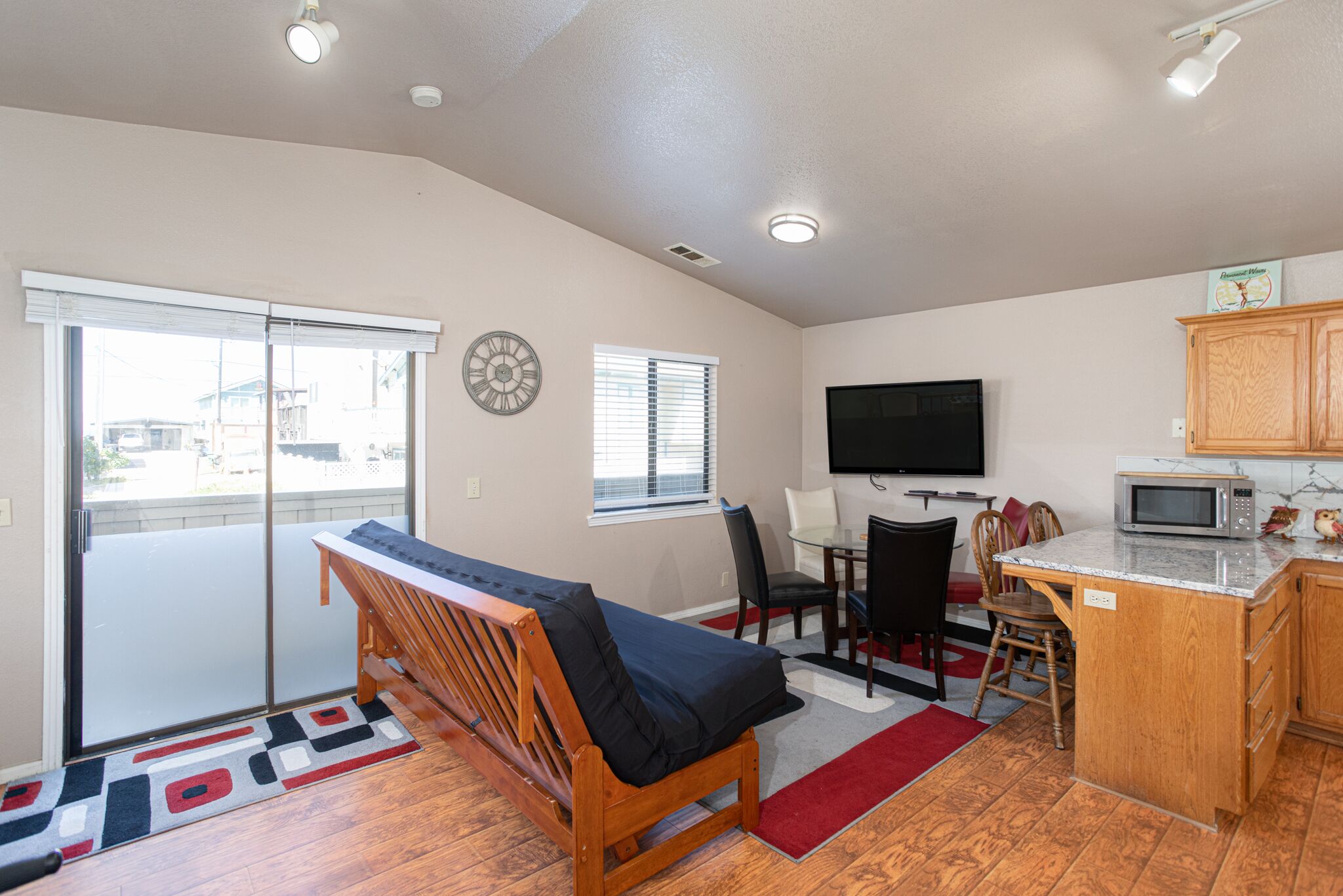 A compact living space with a sliding glass door, a sofa, a dining area with chairs, a kitchen with a microwave, and a flat-screen TV mounted on the wall.