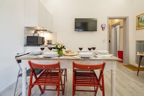 A modern kitchen with a dining table set for two, a flat-screen TV mounted above the table, and a red clock on the wall.