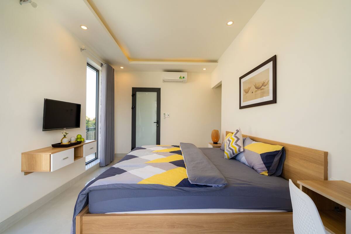 Ein modernes Schlafzimmer mit einem großen Bett, einem Flachbildfernseher, einem Nachttisch aus Holz und einem Fenster mit Vorhängen.