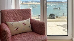 Un sillón rojo con estampado y un cojín, ubicado frente a una puerta corrediza de vidrio que da a un puerto deportivo con barcos y una playa.