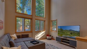 A living room with a large sectional sofa, a flat-screen TV, and multiple windows offering a view of trees and a lake.