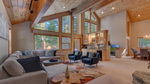 A spacious living area with a large sofa, two armchairs, a coffee table, and a dining table with chairs. There are large windows providing natural light and views of the outdoors.