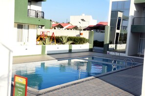 Ein Schwimmbecken mit Hinweisschild, ein Kinderspielbereich und moderne Gebäude im Hintergrund.