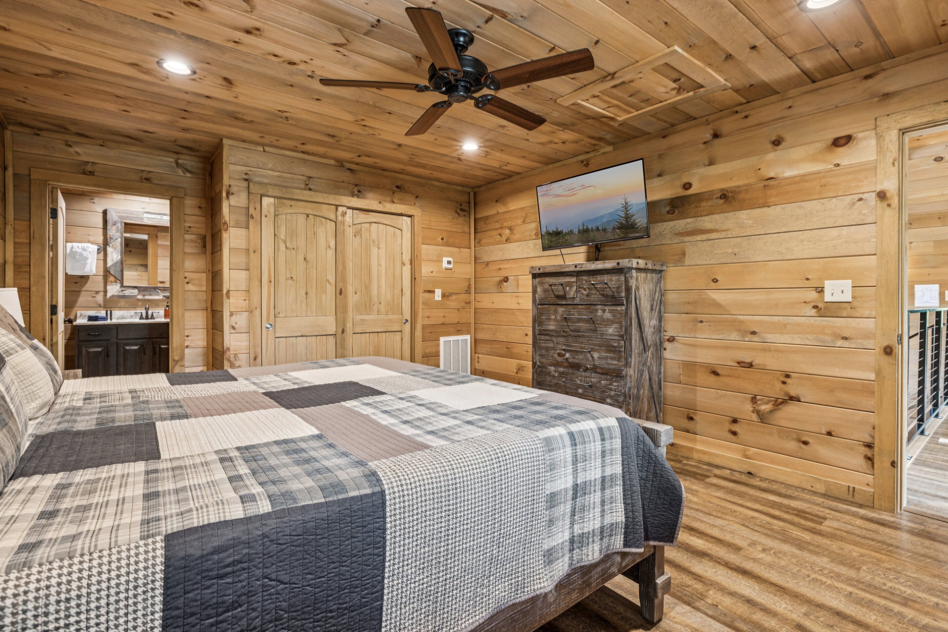 A wooden cabin bedroom with a quilted bedspread, a ceiling fan, and a television on a wooden cabinet.