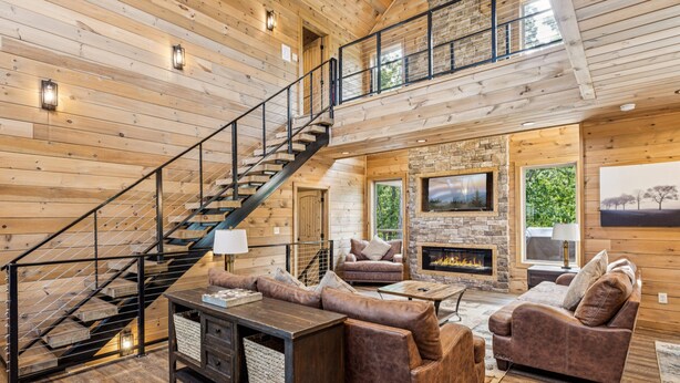 A spacious living area with a stone fireplace, wooden floors, and a staircase with a metal railing.