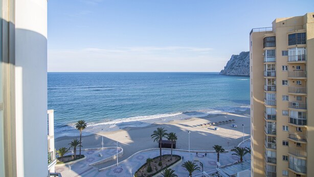 A beach view from a balcony with a clear sky, palm trees, and a rocky cliff in the background. - First line / Wifi / Spa / Seaviews / Heated pool / Pets (Calpe)
