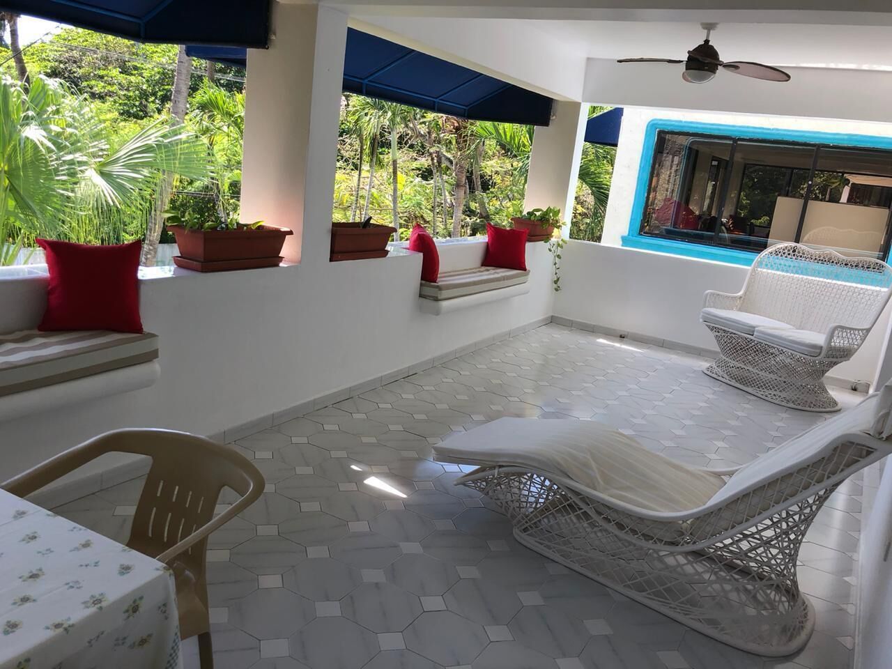 A sunlit patio with hexagonal tile flooring, outdoor seating with cushions, potted plants, and a view of lush greenery.