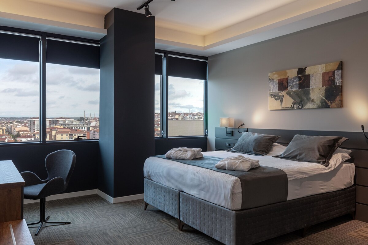 Quarto moderno com uma cama grande, uma escrivaninha e uma cadeira. Apresenta vista para a cidade através de grandes janelas.
