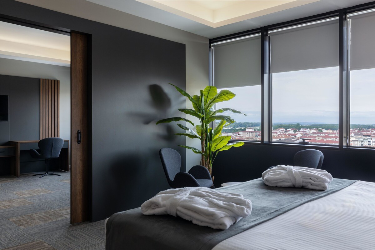 Quarto de hotel moderno com uma cama grande, uma escrivaninha, uma cadeira e vista da cidade através de grandes janelas.