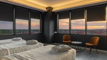 Ein Hotelzimmer mit zwei Betten, einem kleinen Tisch und Stühlen, großen Fenstern mit Blick auf die Stadt.