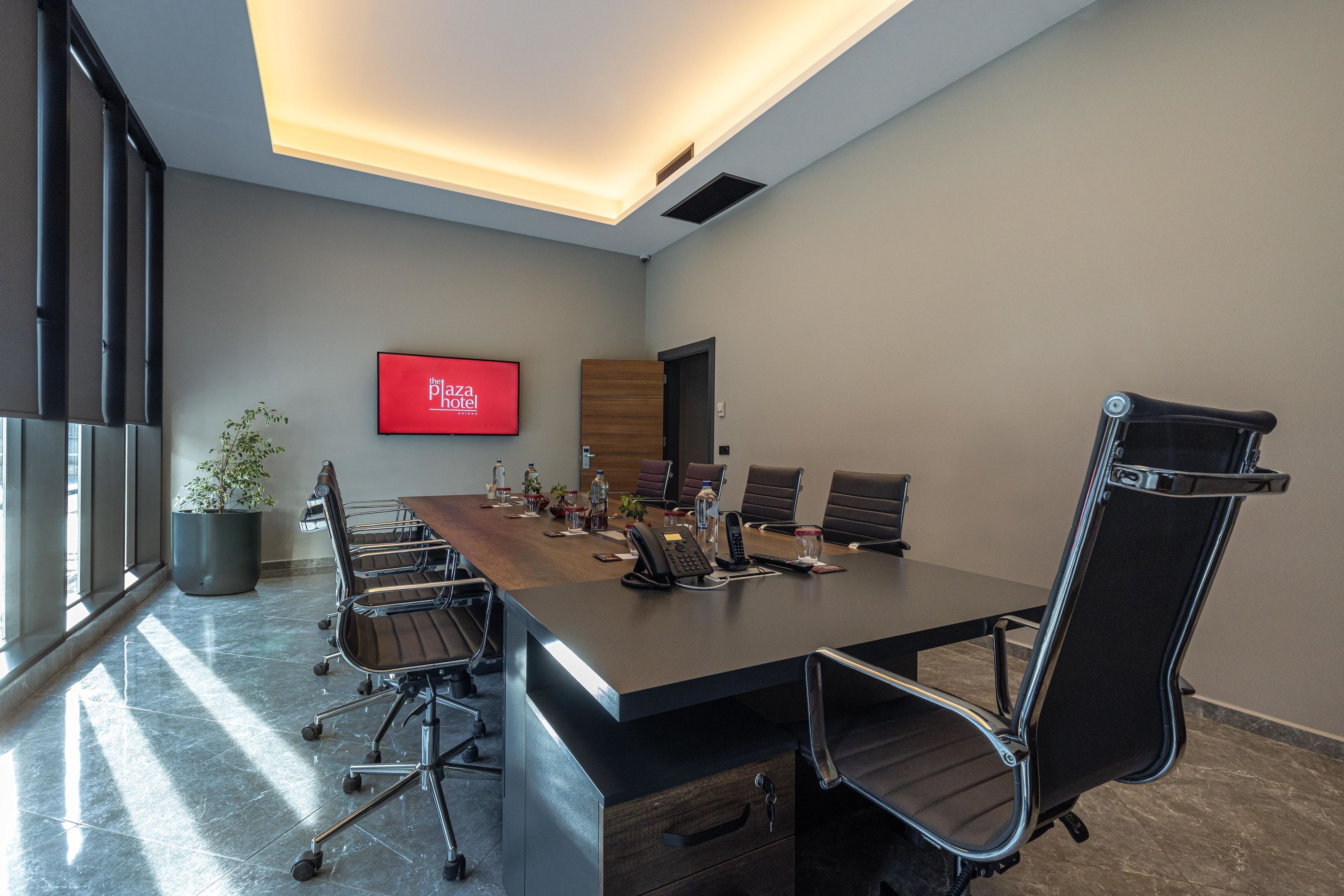 Uma sala de conferências com uma mesa comprida, cadeiras, uma televisão e um vaso de planta.
