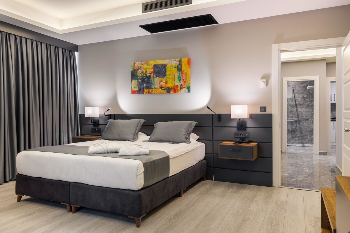 Quarto moderno com uma cama grande, duas mesas de cabeceira, um quadro abstrato na parede e vista para o banheiro adjacente.