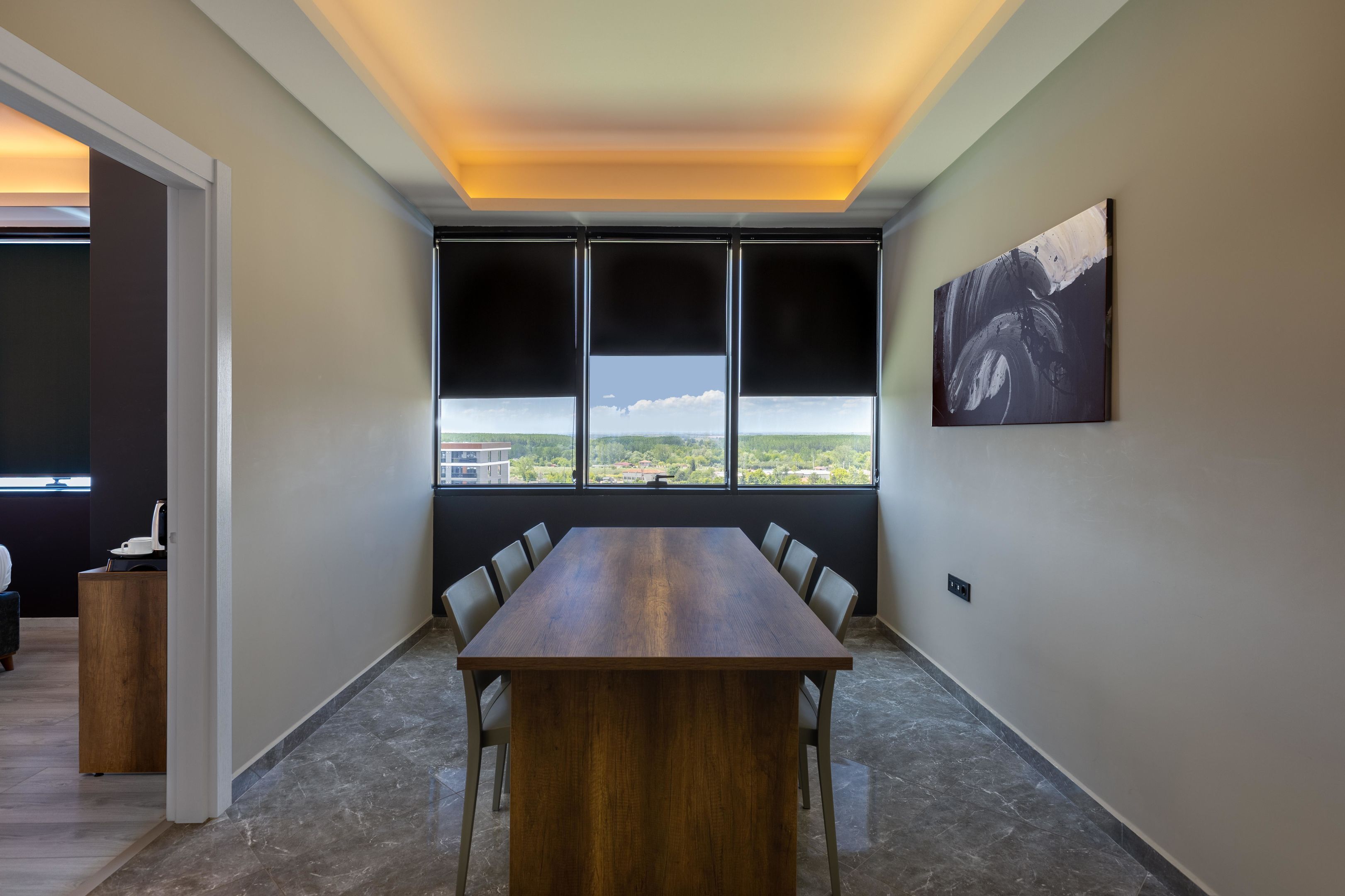 A conference room with a long wooden table, multiple chairs, a large window with a view, and a painting on the wall.