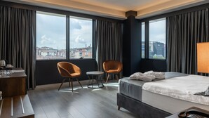 Ein Hotelzimmer mit einem großen Bett, zwei Sesseln, einem kleinen runden Tisch, einem Schreibtisch mit Lampe und Blick auf die Stadt durch große Fenster.