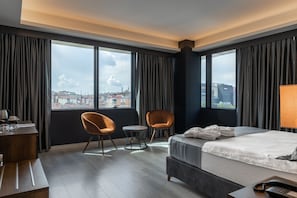 A hotel room with a large bed, two armchairs, a small round table, a desk with a lamp, and a view of the city through large windows. - The Plaza Hotel Edirne (Edirne)