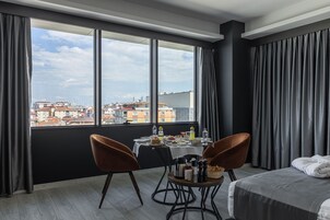 A hotel room with a large window, a bed, two chairs, a table with breakfast items, and a view of the cityscape.