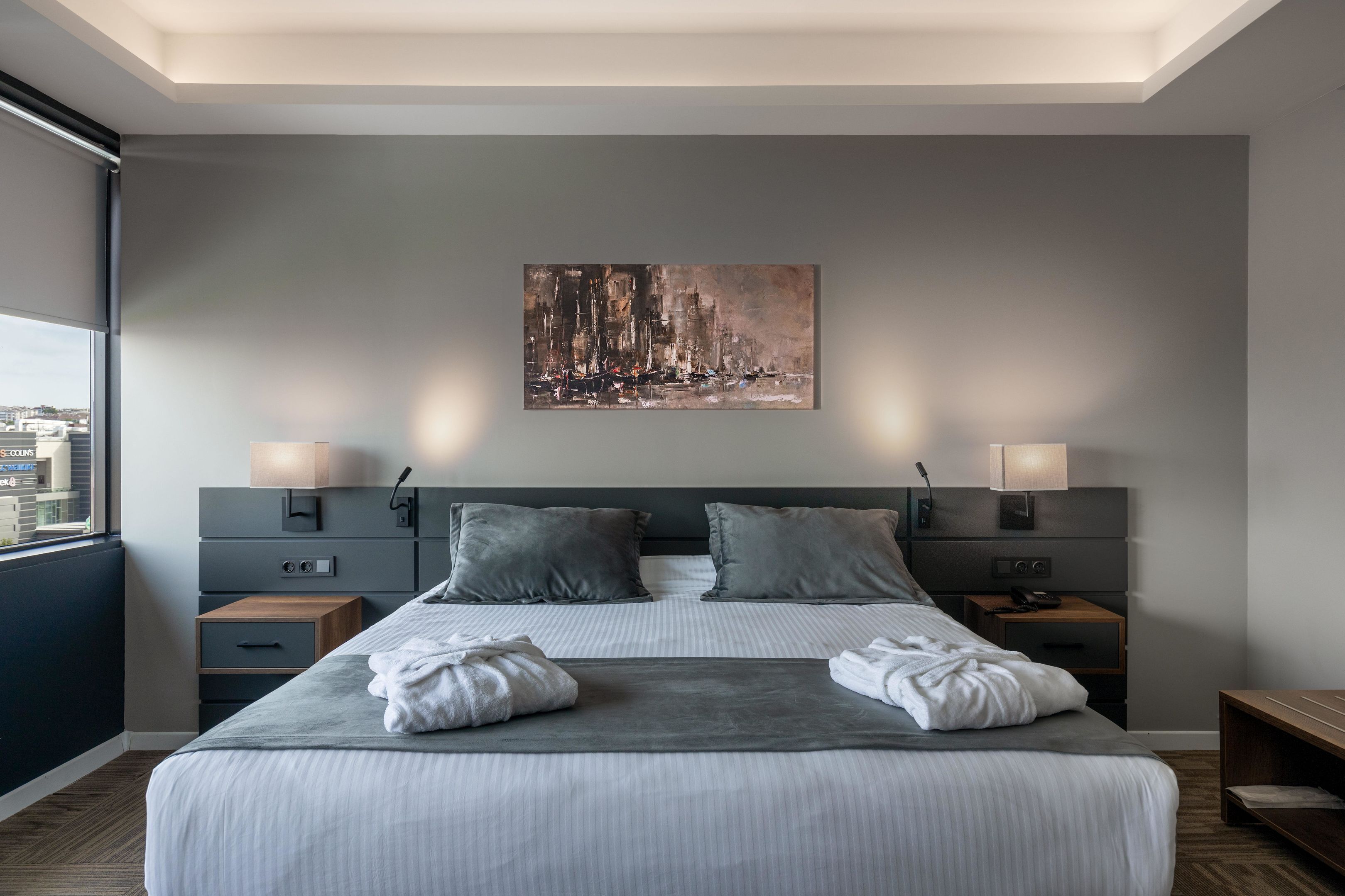 Une chambre d’hôtel moderne avec un grand lit, deux tables de chevet, un tableau au mur et une vue sur la ville par la fenêtre.