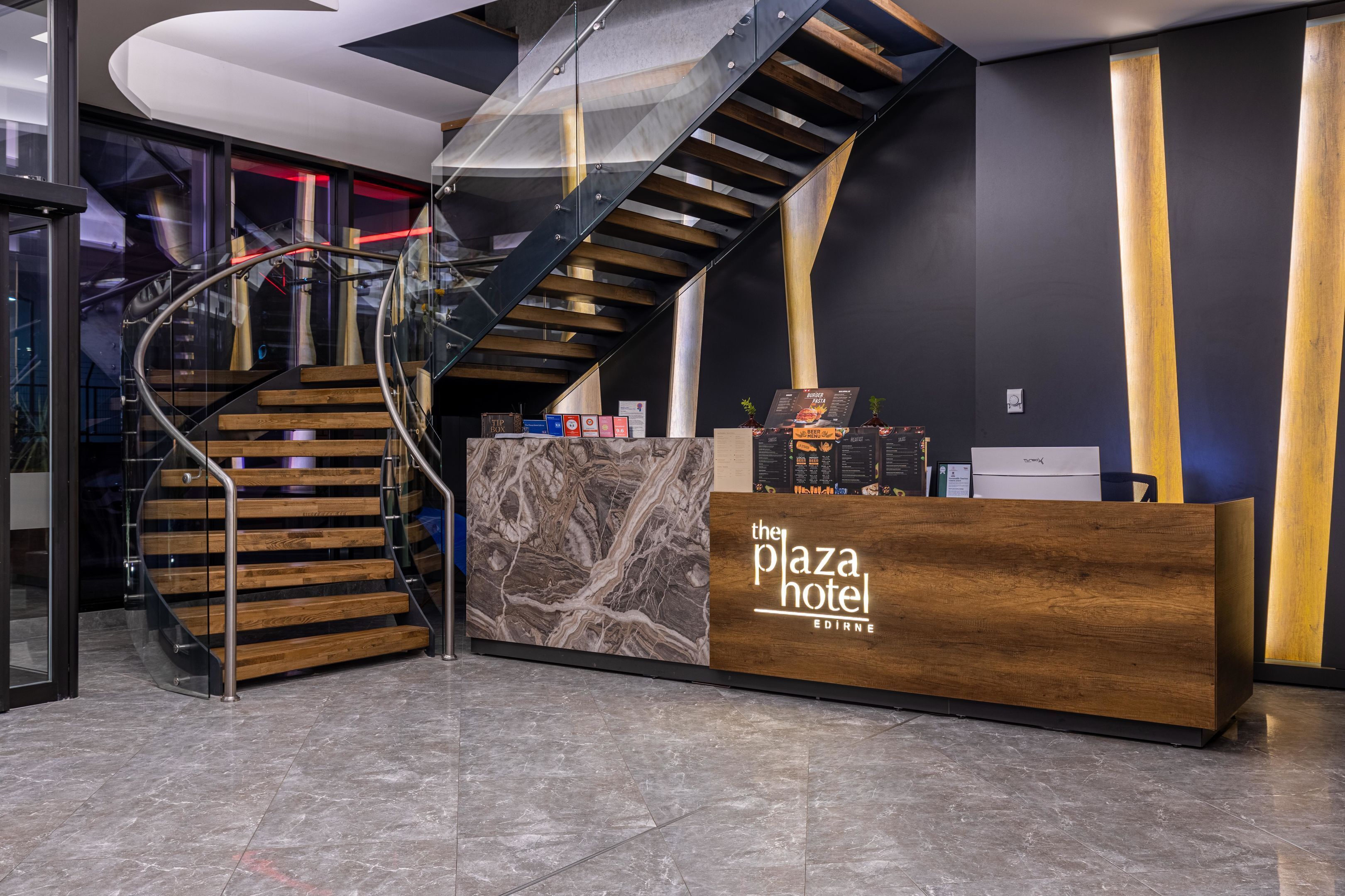 Een receptiebalie met een houten afwerking en een marmeren accent, een trap met houten treden en metalen leuningen, en een modern interieur met donkere muren en een gebogen plafond.