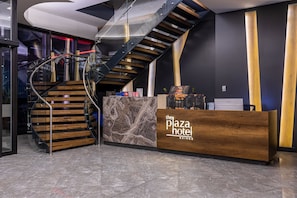 A reception desk with a wooden finish and a marble accent, a staircase with wooden steps and metal railings, and a modern interior design with dark walls and a curved ceiling.