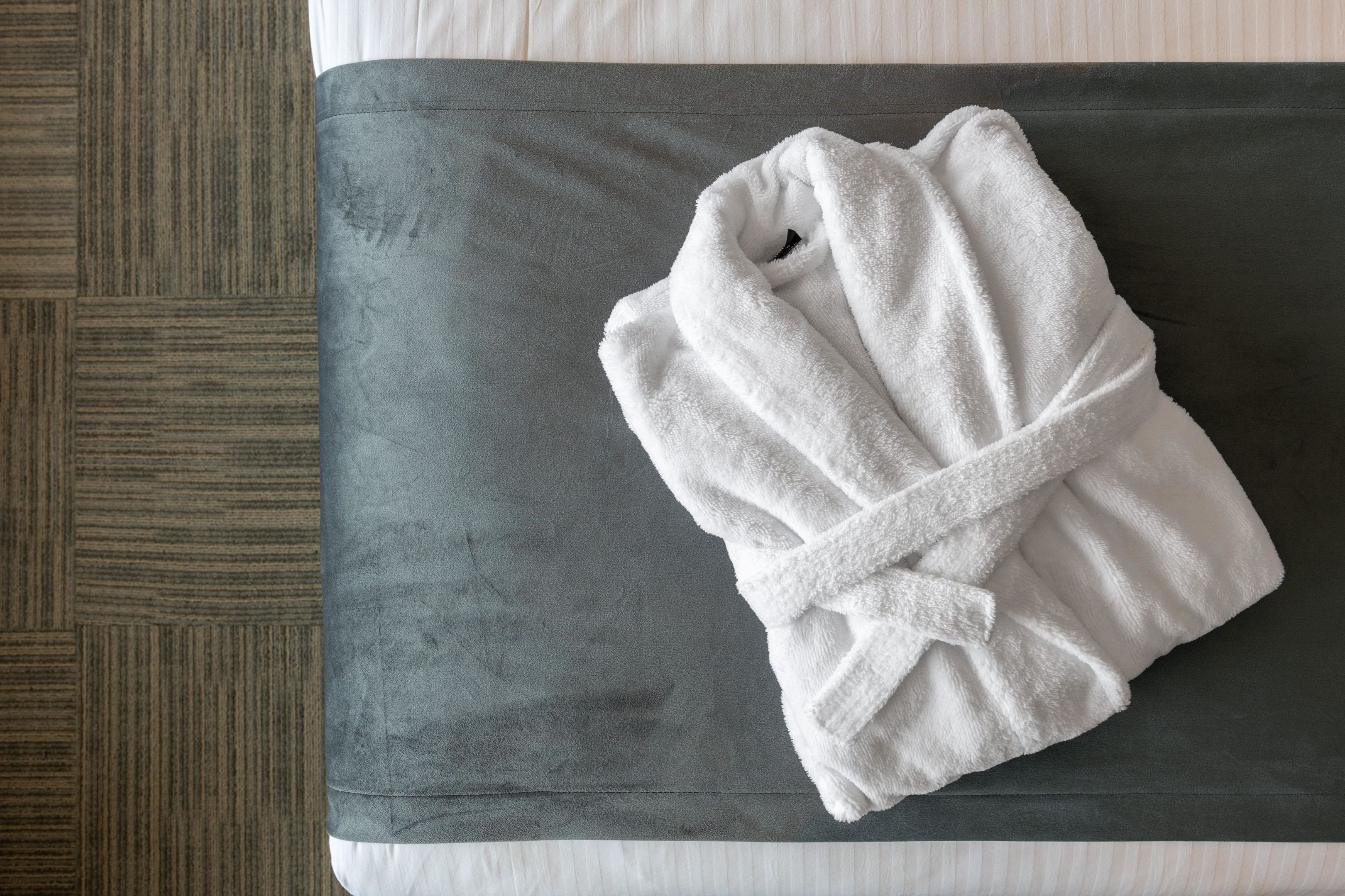 A neatly folded white bathrobe on a grey cushioned bench beside a bed with white bedding.