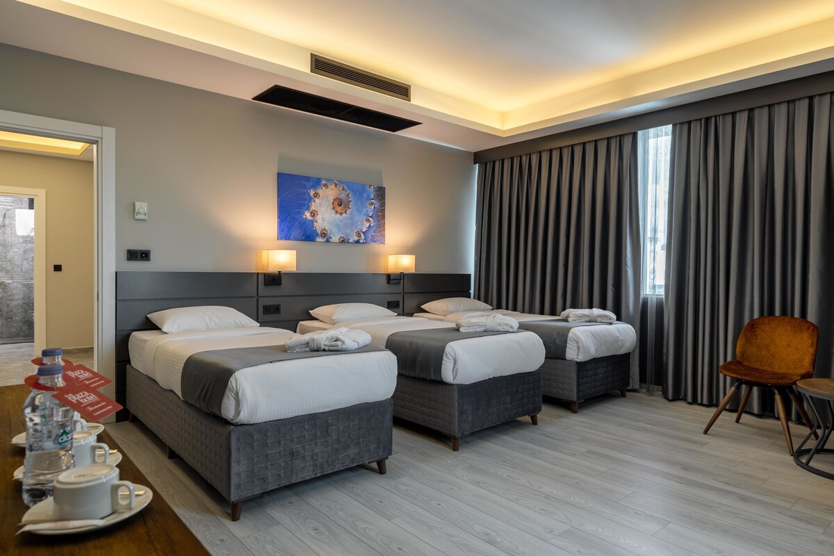 Quarto de hotel com três camas, uma mesa de jantar com itens de café da manhã, um quadro na parede e uma janela grande com cortinas.