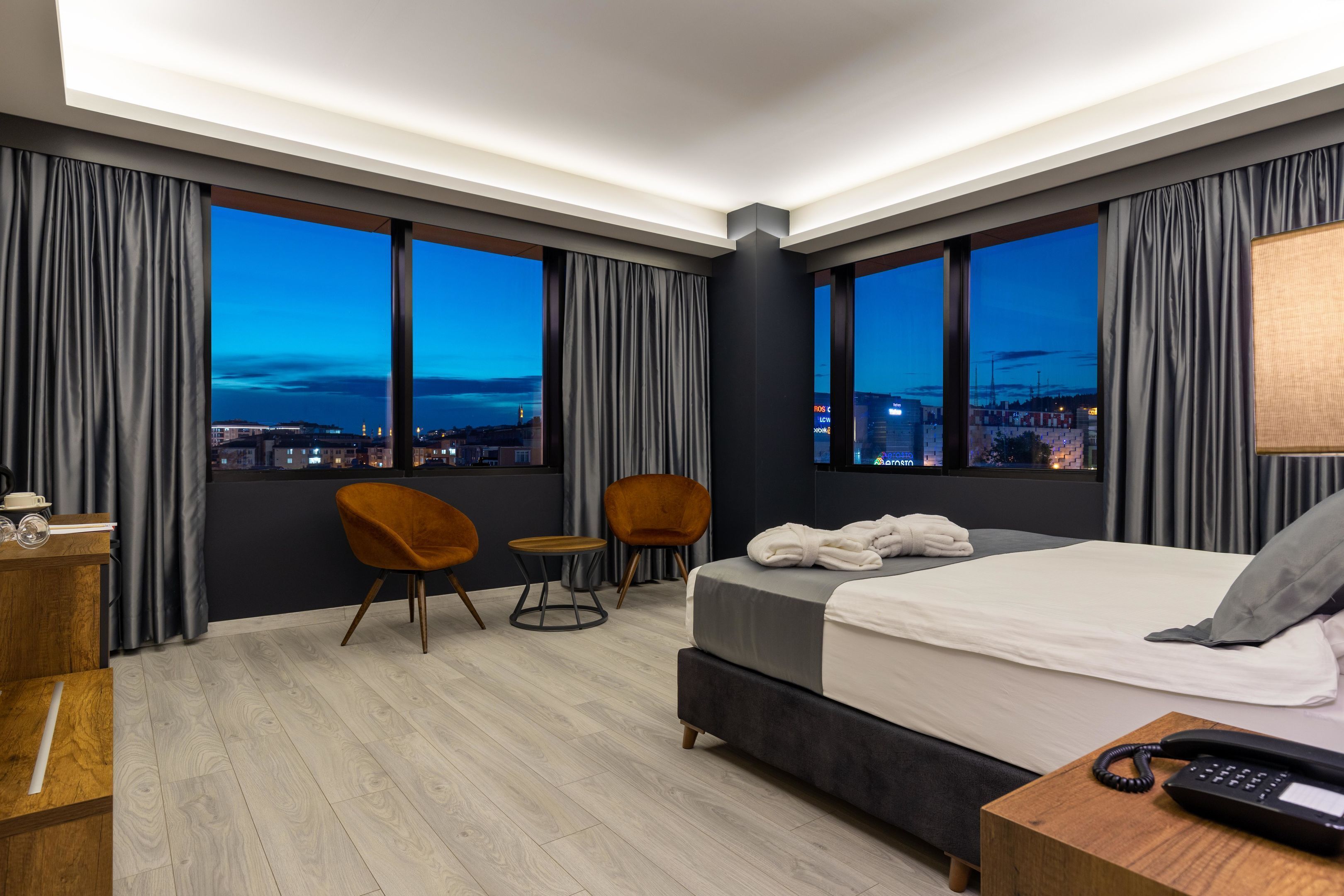 A modern hotel room with a large bed, two chairs, a desk, and a view of the city at dusk.