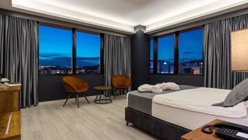 A modern hotel room with a large bed, two chairs, a desk, and a view of the city at dusk.